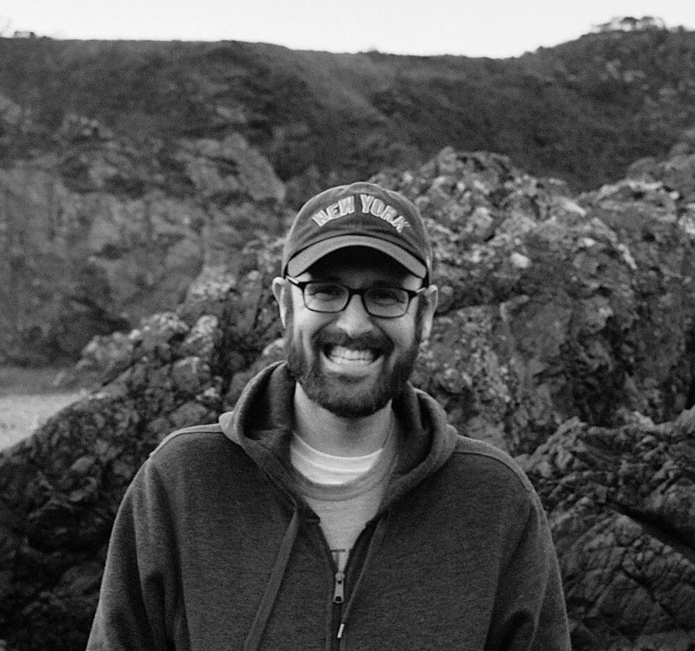 Josh Catone, smiling, in a New York cap in front of a rocky hillside